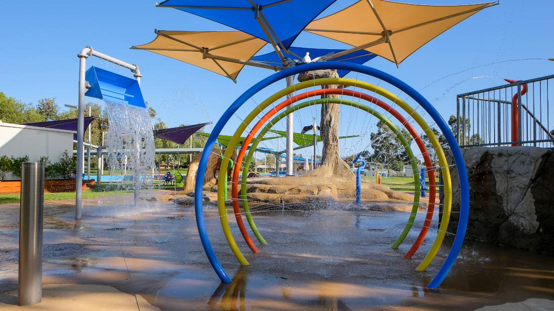 Colourful water play area with rainbow arch and shade umbrellas in park.