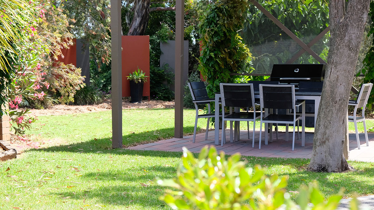 Covered BBQ and outdoor dining area with lawn and flowering shrubs.